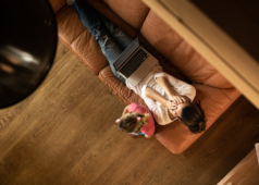 A stay-at-home mom lying on a couch with her child nearby, looking concerned while browsing on a laptop.